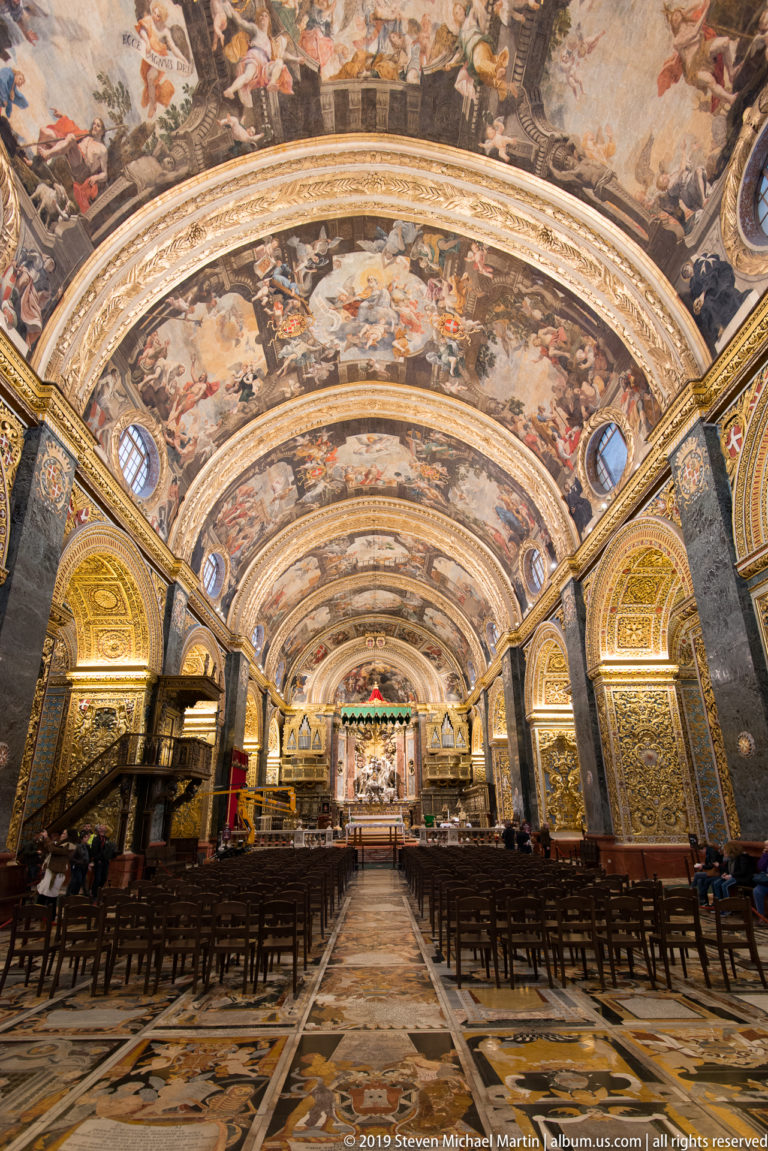 Low centered view down the nave with memorial pavement, rows of chairs, gilded arches, and painted barrel vault.
