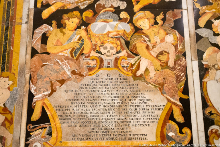 Inlaid marble tomb slab with two angels, skull, drapery, Latin inscription, and colorful floral stonework.