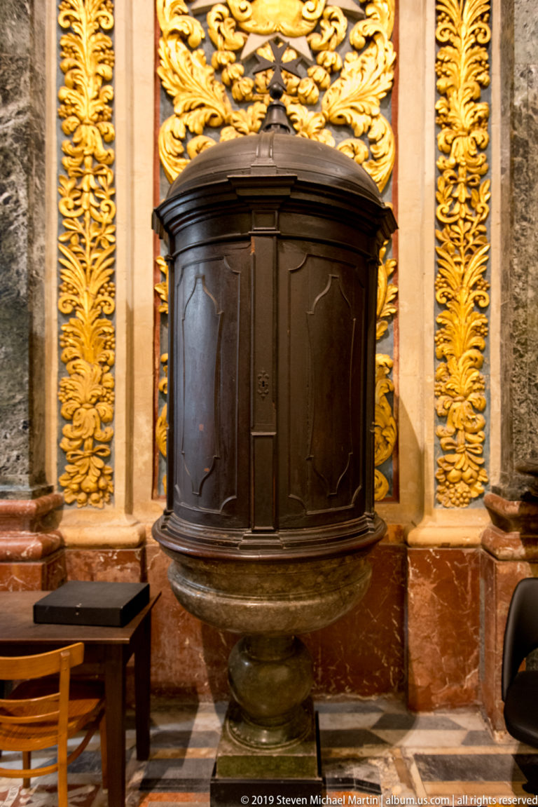 Dark polygonal wooden enclosure mounted against gilded wall panels and colored marble in the cathedral interior.