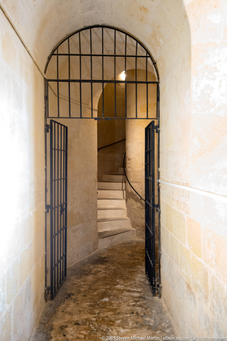 Plain limestone corridor with open black iron gates leading to pale stone steps beneath a rounded arch.