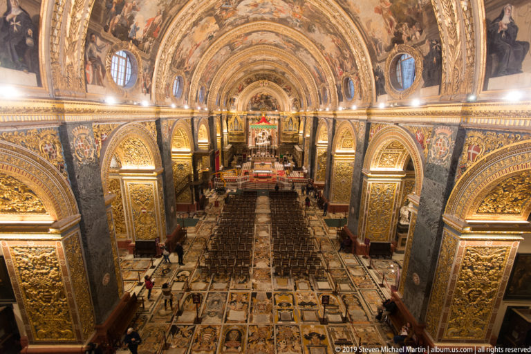 High axial view down the nave, showing memorial pavement, rows of chairs, gilded arches, and painted barrel vault.