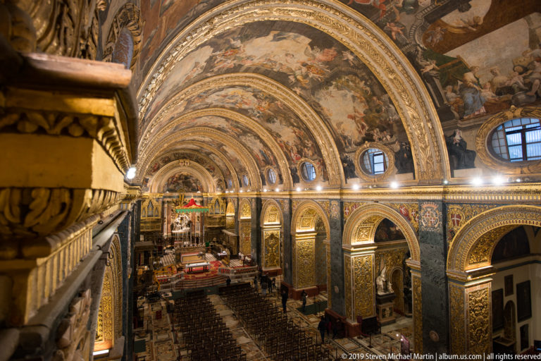 Elevated side view along the cathedral nave beneath a painted barrel vault, with chairs, marble floor, and gilded arches.