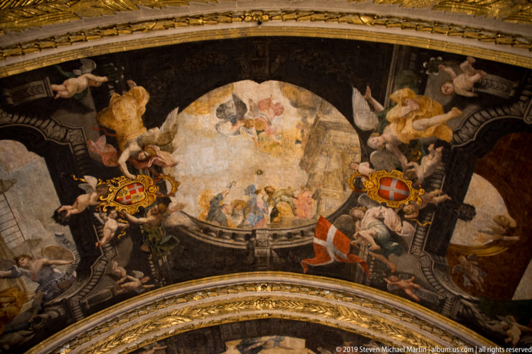 Curved painted vault with angels, central fresco, dark architectural framing, and colorful heraldic shields.