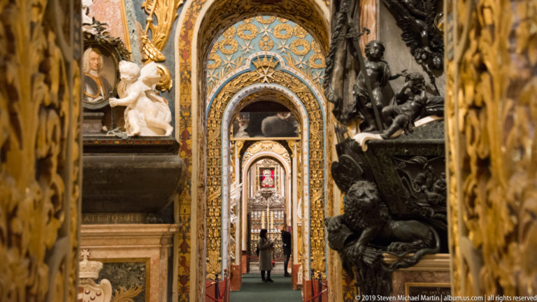 Aligned ornate chapel doorways with blue-and-gold decoration, sculptural monuments, and a distant gated opening.