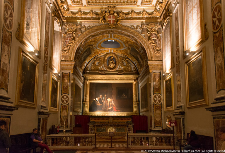 Ornate chapel with large altarpiece, gilded arch, marble details, balustrade, and visitors at the sides.
