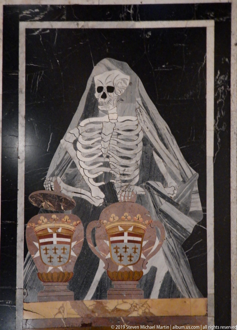 Inlaid black marble tomb slab showing a veiled skeleton above two colored heraldic shields.