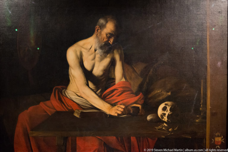 Elderly Saint Jerome in red drapery writing at a table beside a skull against a dark background.