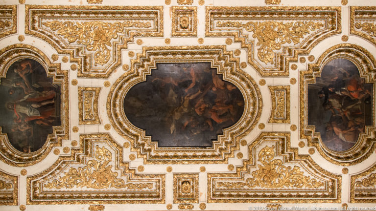 Dark central ceiling painting enclosed by stepped gilded moldings, foliate relief, and smaller framed panels.