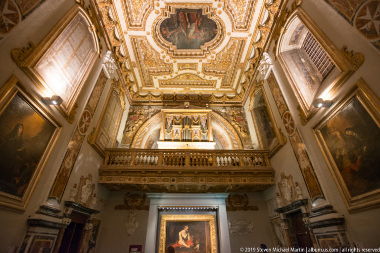 Tall chapel interior with altar painting, projecting gallery, high windows, framed paintings, and ornate painted ceiling.
