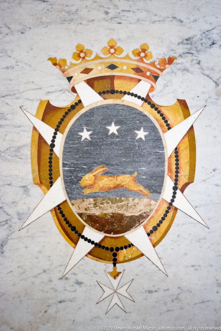Crowned circular heraldic emblem inlaid in marble, with two white stars, radiating points, and colored stone borders.