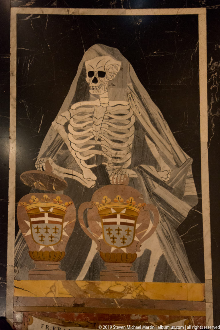 Inlaid marble skeleton wrapped in a shroud above two crowned heraldic emblems on a dark floor panel.