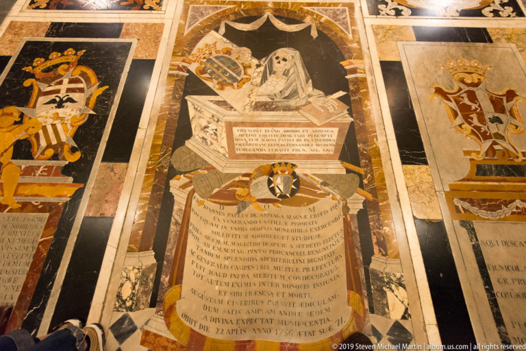 Inlaid marble tomb slab with robed effigy, Latin inscription, heraldic emblems, and contrasting stone borders.