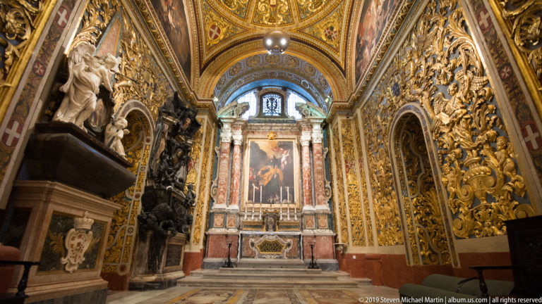 Side chapel altar with painted altarpiece, colored marble columns, gilded relief walls, and sculptural monuments.