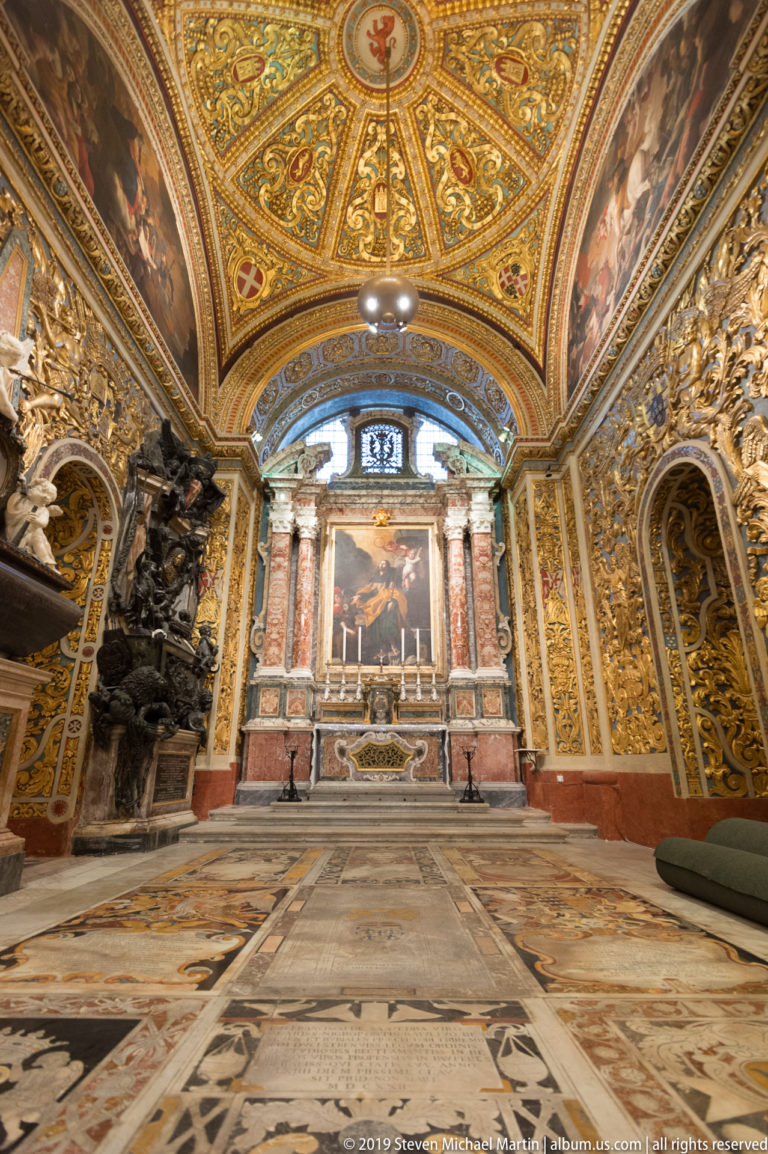 Ornate side chapel with patterned marble floor, gilded walls, monuments, columns, and a painted altarpiece.