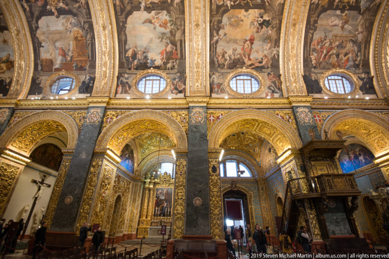 Cathedral interior with gilded arcade, dark marble piers, oval windows, painted vault scenes, visitors, and wooden pulpit.