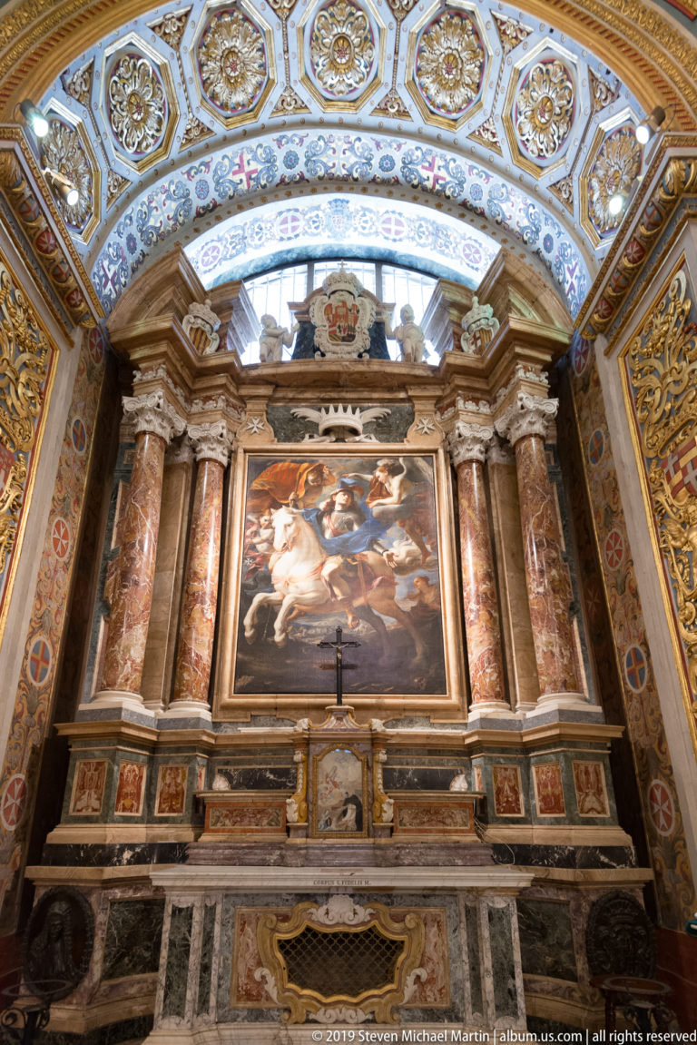 Large religious painting framed by red marble columns above a polychrome marble altar wall and blue-and-gold patterned ceiling.