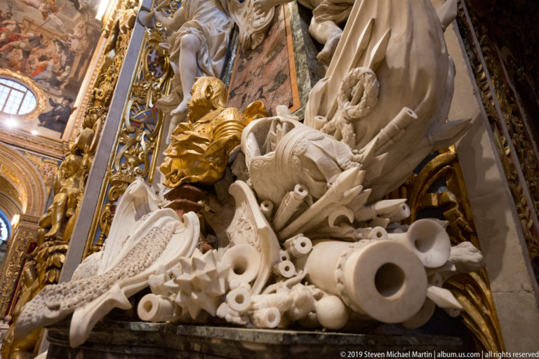 Close marble carving of cannon barrels, shields, helmets, standards, and other military trophies beneath a gilded figure.