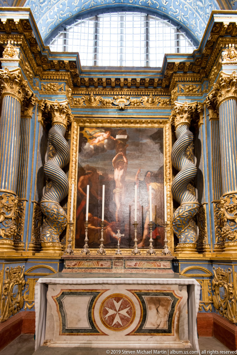 Side chapel altar with central painting, blue-and-gold twisted columns, high window, candles, and Maltese cross on altar frontal.