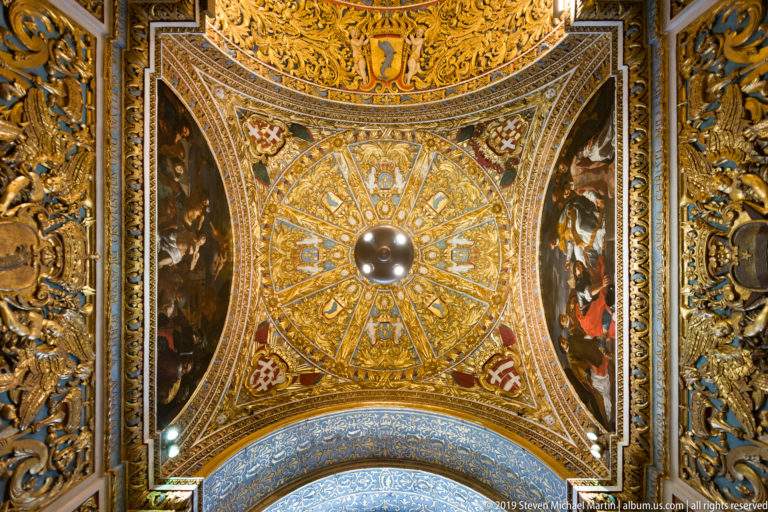 Upward view of radial gilded ceiling decoration with blue panels, central boss, heraldic crosses, and adjacent painted scenes.