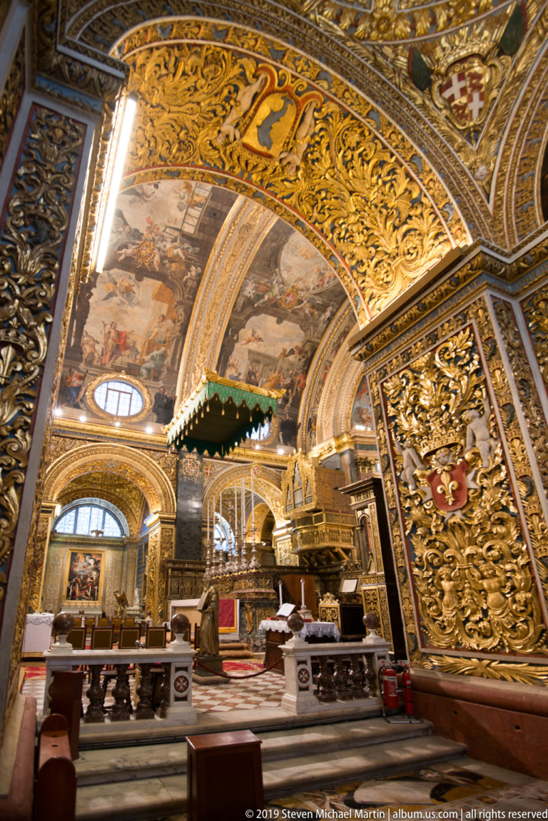 Oblique side-aisle view with painted vaults, gilded arches, chapel openings, seating, and dark elevated wooden furnishing.