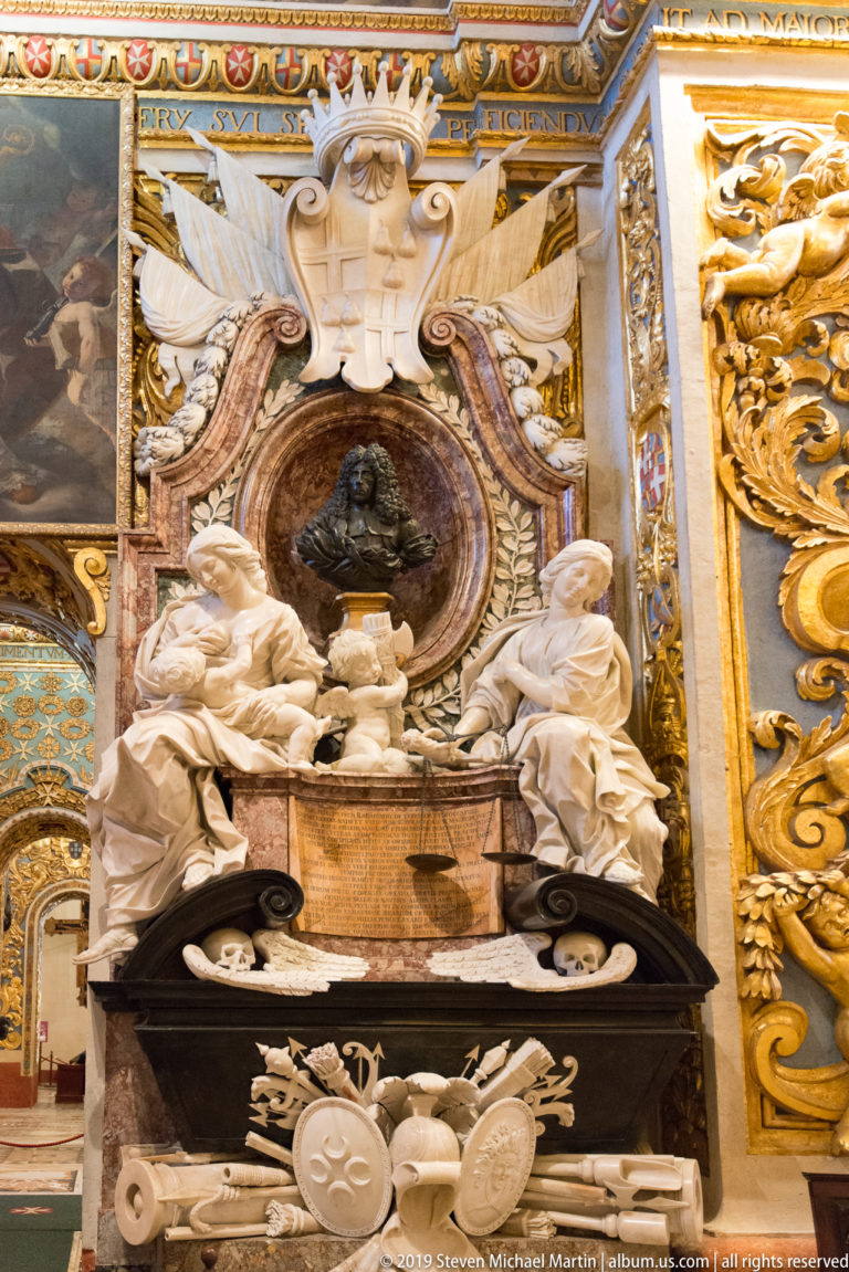 Marble funerary monument with reclining figures, dark portrait medallion, and carved helmets, shields, cannon barrels, and standards.