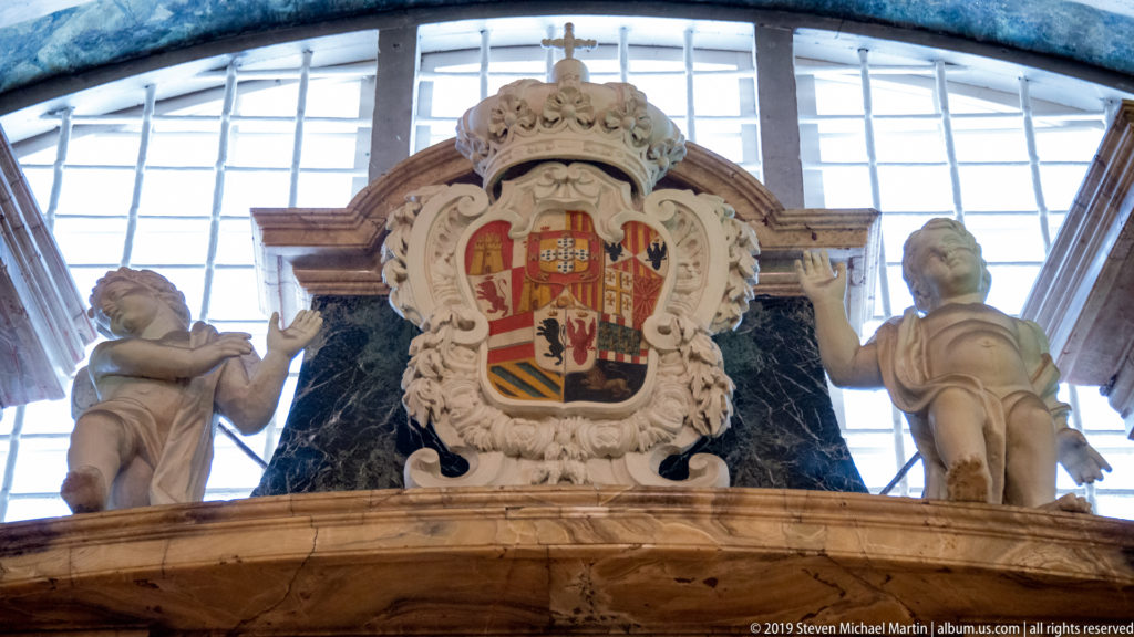Large coat of arms on dark marble monument flanked by two pale statues beneath a bright window.