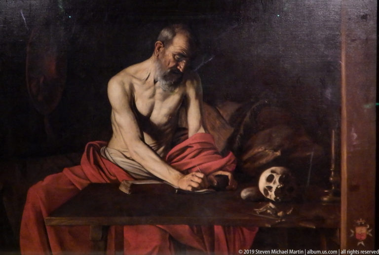 Caravaggio’s Saint Jerome writes at a dark table beside a skull, his bare torso and red drapery illuminated against deep shadow.