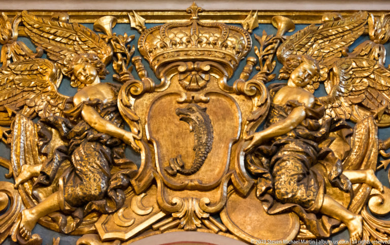 Gilded heraldic shield flanked by winged putti and surrounded by dense scrolling relief ornament.