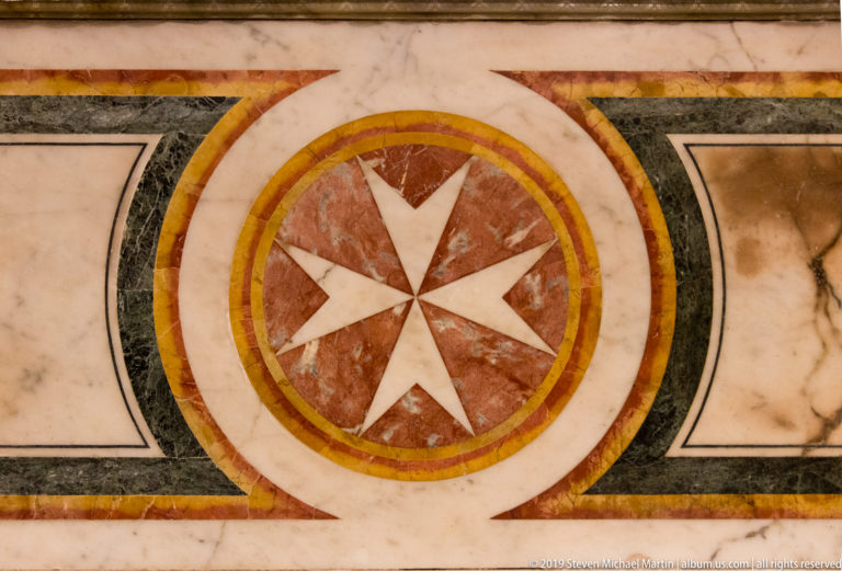 Eight-pointed white Maltese cross in a circular red, gold, green, and pale marble floor inlay.