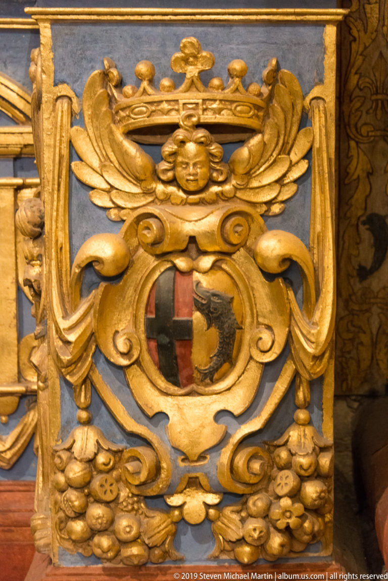 Crowned gilded cartouche around a quartered shield with red cross and dark animal device, framed by cherub and fruit.