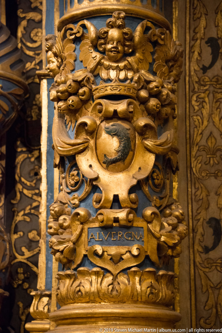 Close gilded heraldic shield labeled “ALVERGNA,” crowned by a cherub and surrounded by scrolling foliage.