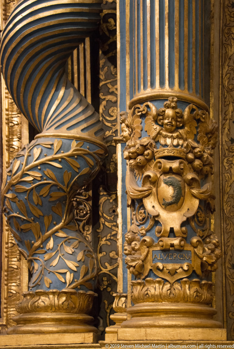 Gilded shield with cherub and “ALVERGNA” inscription on a blue-and-gold pilaster beside a twisted column.