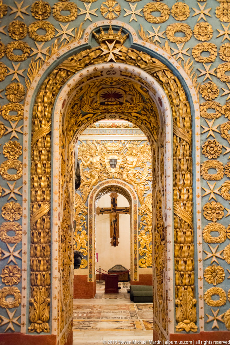 Series of gilded rounded arches with blue patterned panels leading toward a crucifix in a side passage.