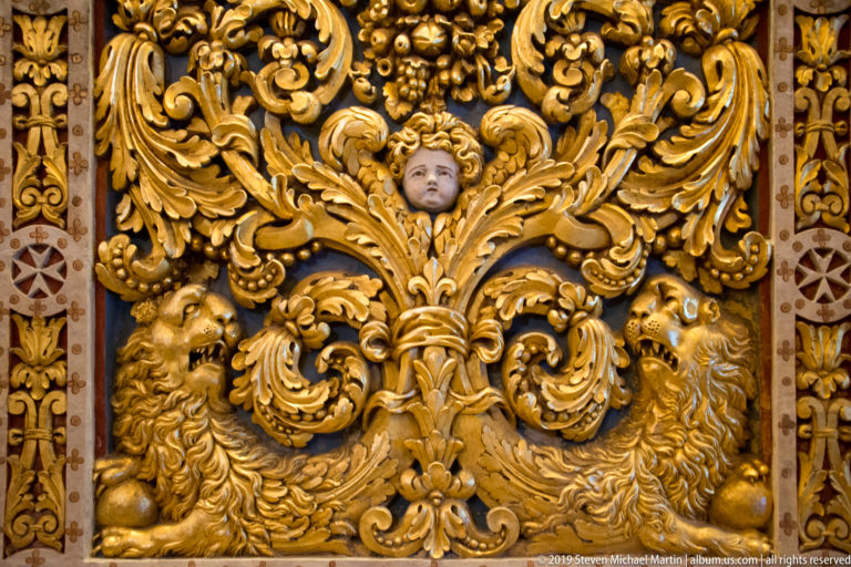 Gilded relief panel with central cherub, curling foliage, fruit, and lion heads at the lower corners.