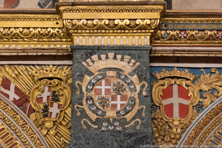 Three heraldic emblems set among polychrome marble panels, gilded scrollwork, and patterned architectural bands.