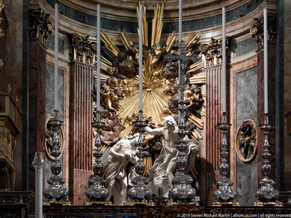 Christ and John the Baptist stand in a sculptural Baptism group before gilded rays, surrounded by angels, clouds, and altar furnishings.