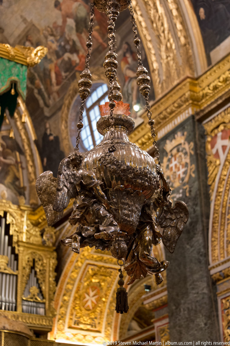 Close view of an ornate metal hanging lamp suspended by chains against gilded and painted cathedral decoration.