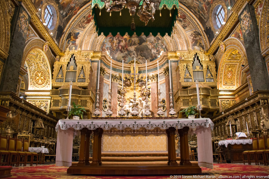 Frontal high altar with tall candles, paired organ cases, choir stalls, gilded walls, and painted vaulting.