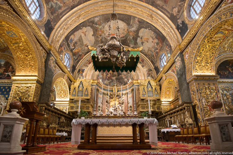High altar centered beneath a painted and gilded vault, with organ cases, choir stalls, and a large hanging lamp.