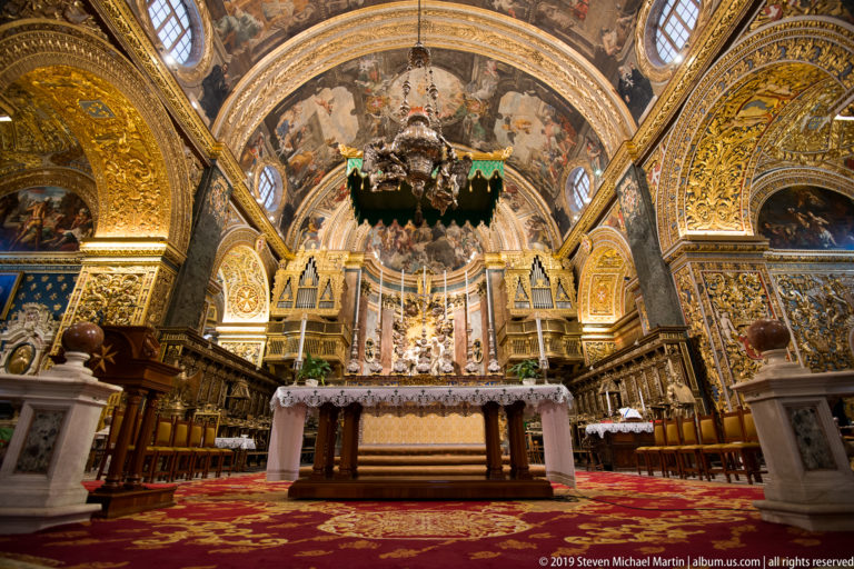 Wide low-angle sanctuary view with high altar, hanging lamp, organ cases, choir stalls, and gilded painted vaults.