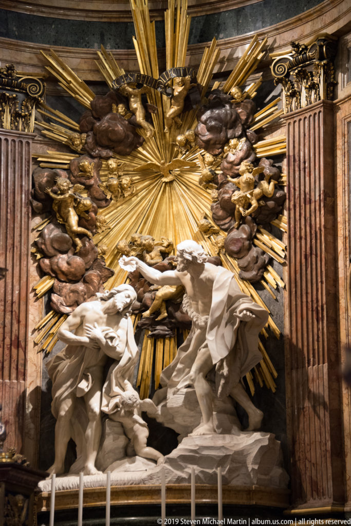 White sculpted figures of Christ and John the Baptist stand before gilded rays, surrounded by angels and dark clouds.