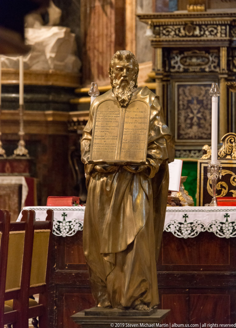 Metal figural lectern holding an open book, with altar furnishings, candles, and polychrome decoration behind.