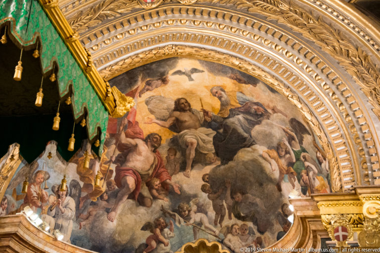 Religious ceiling painting of figures among clouds framed by a broad gilded arch, with a green canopy at left.