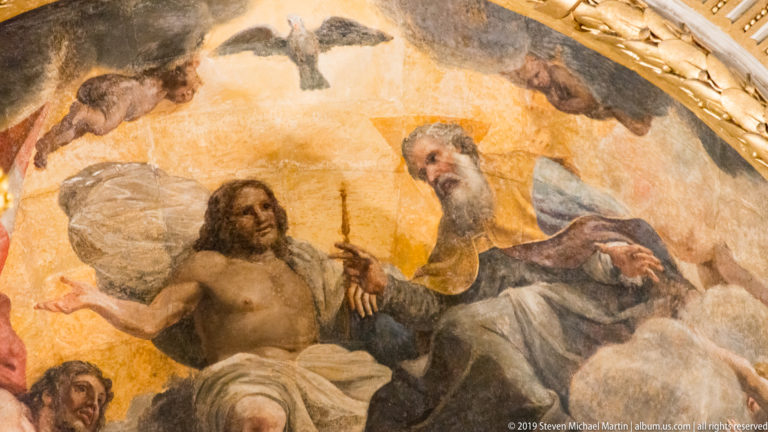 Close ceiling-painting detail of a seated draped man beside an elderly bearded figure holding a slender rod amid clouds.