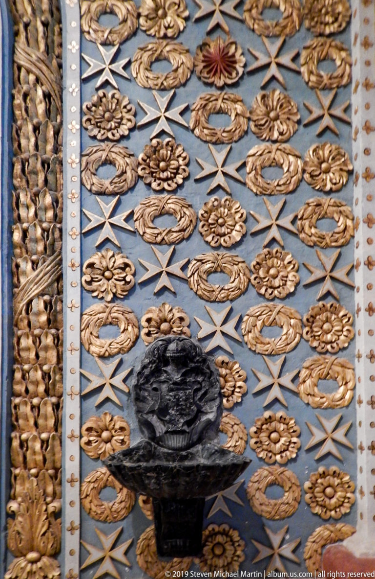 Dark bust mounted on a blue wall patterned with gilded wreaths, rosettes, and repeated Maltese crosses.