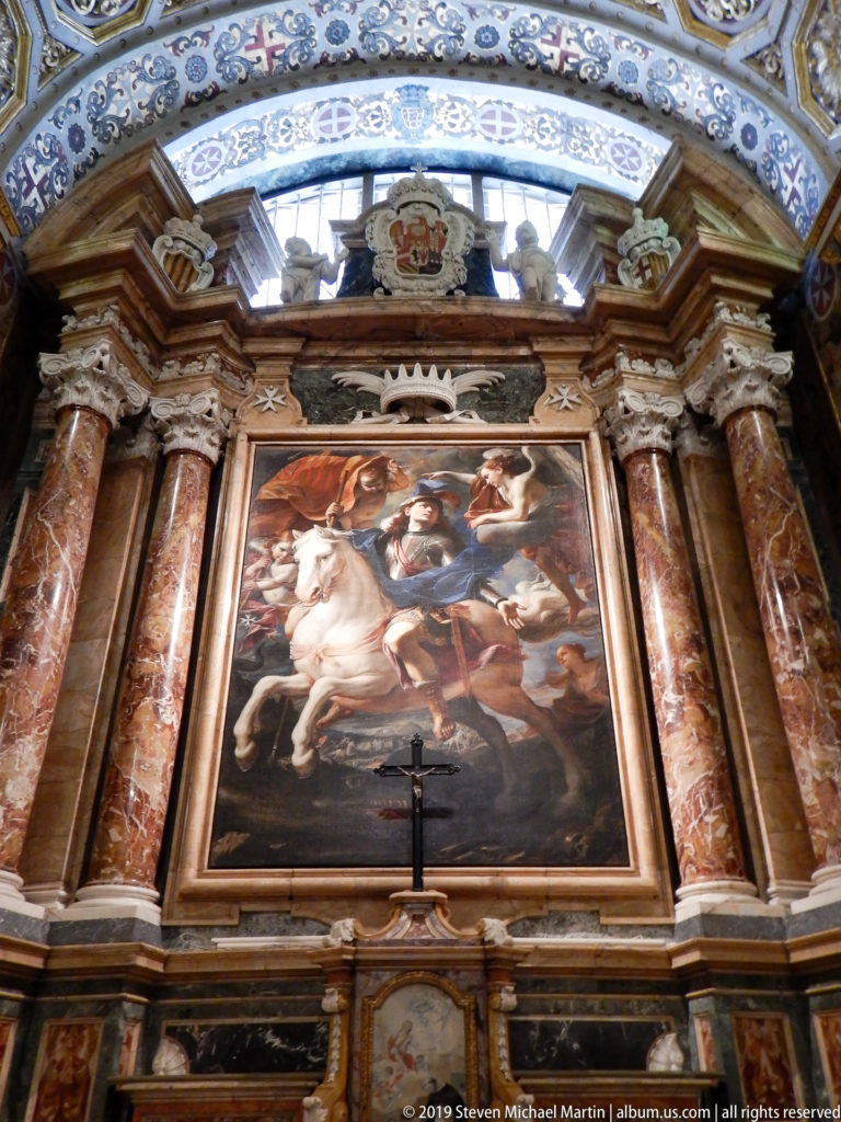 Large religious painting above an altar, framed by reddish marble columns and elaborate architectural decoration.