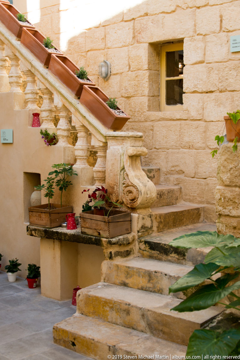 Short weathered limestone staircase with carved scroll balustrade, potted flowers, and plants beside a honey-colored stone wall.