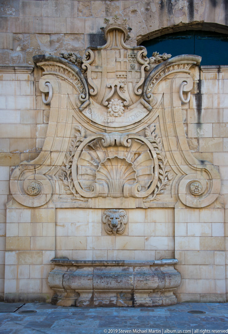 Ornate limestone wall fountain with large scrolls, shell motifs, central outlet, heraldic carving, and a projecting basin.
