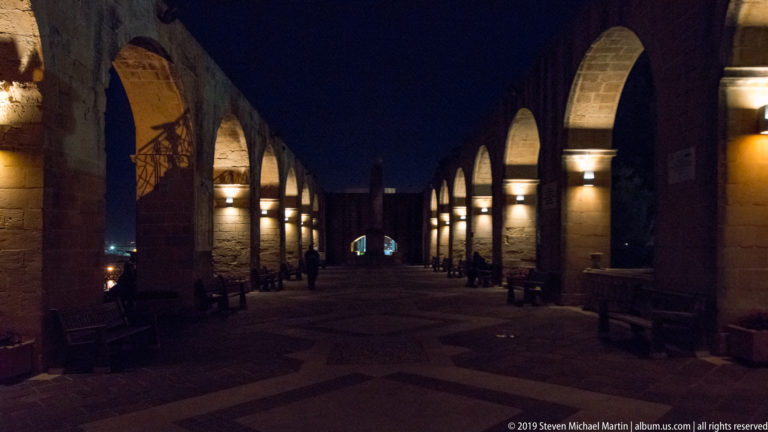 Long nighttime stone arcade with repeated arches, wall lights, benches, and a few pedestrians receding into the distance.