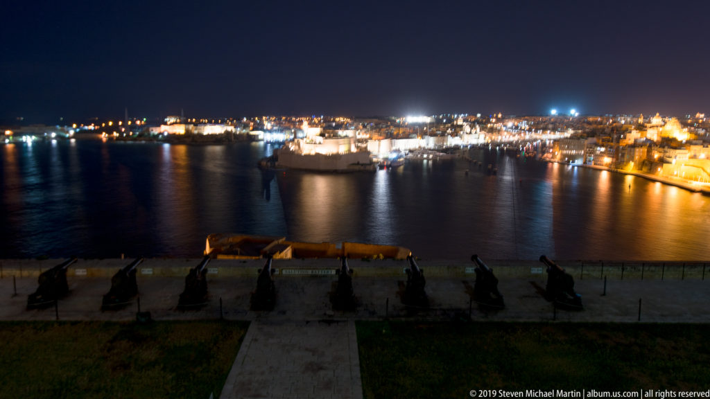 Wide night view over a harbor with illuminated shorelines, a large vessel on the water, and a dark terrace in the foreground.